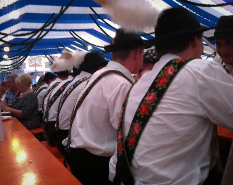 At least six young men sit side to side in a German fest tent, all wearing traditional suspenders. Not So SAHM